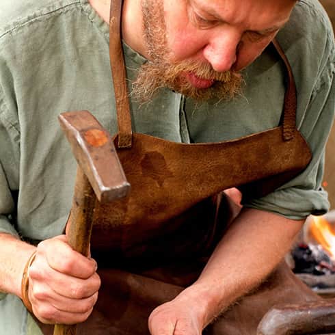 The image shows a bearded blacksmith forging a socket for an arrowhead, beating hte hot metal with his hammer.