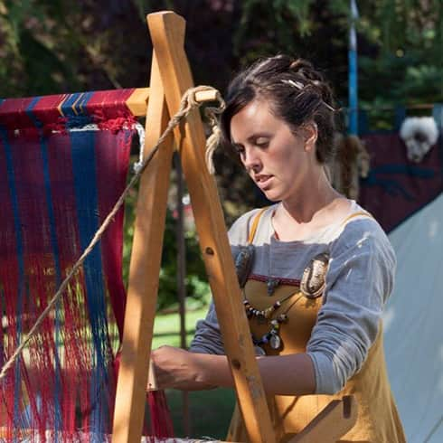 The image shows a woman weaving woollen fabric on a warp-weighted loom.