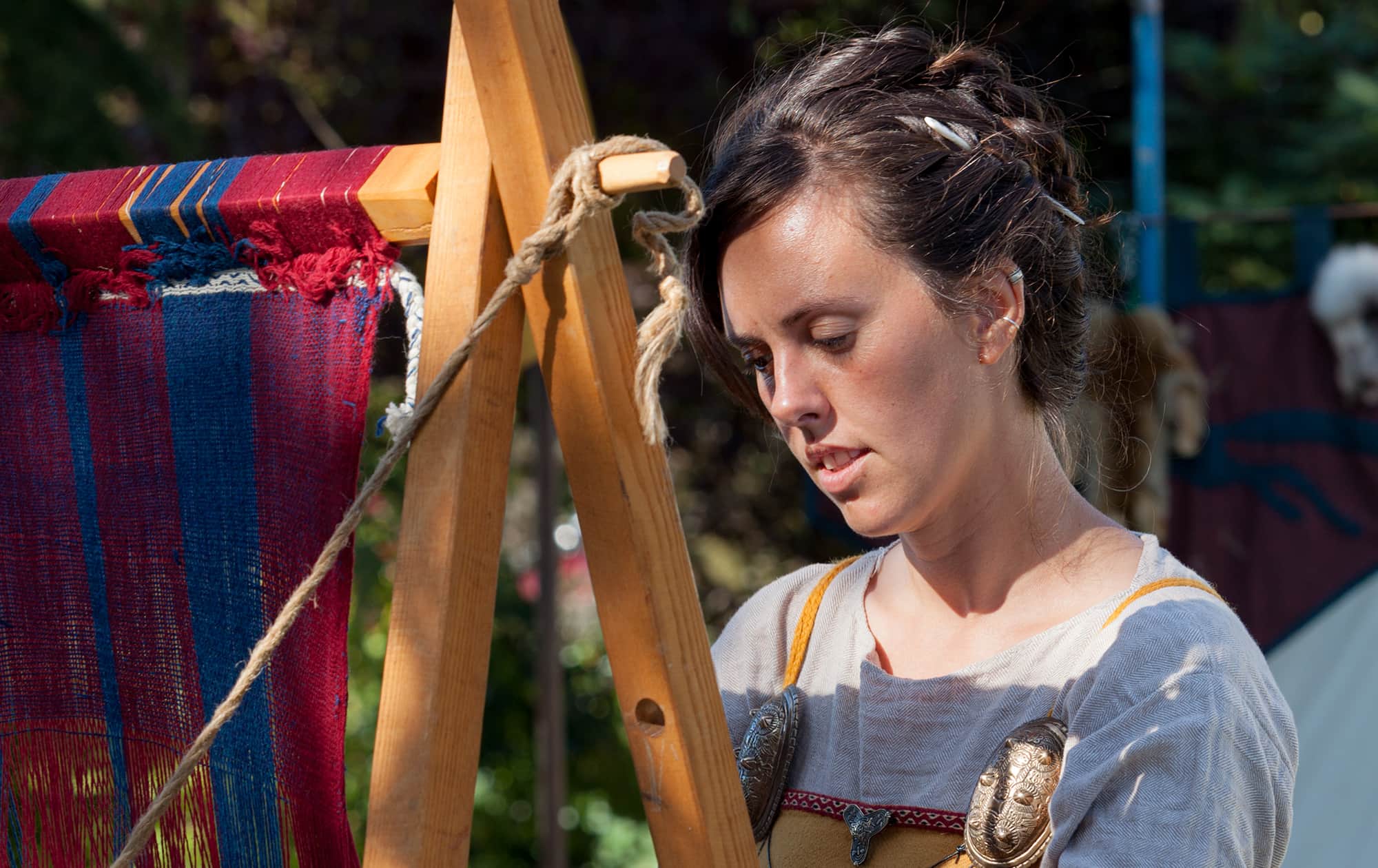 Gytha the weaver passed the shuttle of the weft yarn throup the warp in her weave. She uses a warp-weighted loom with red and blue yarn to weave her woollen fabric. She wears a mustard suspended dress pinned with bronze oval brooches.